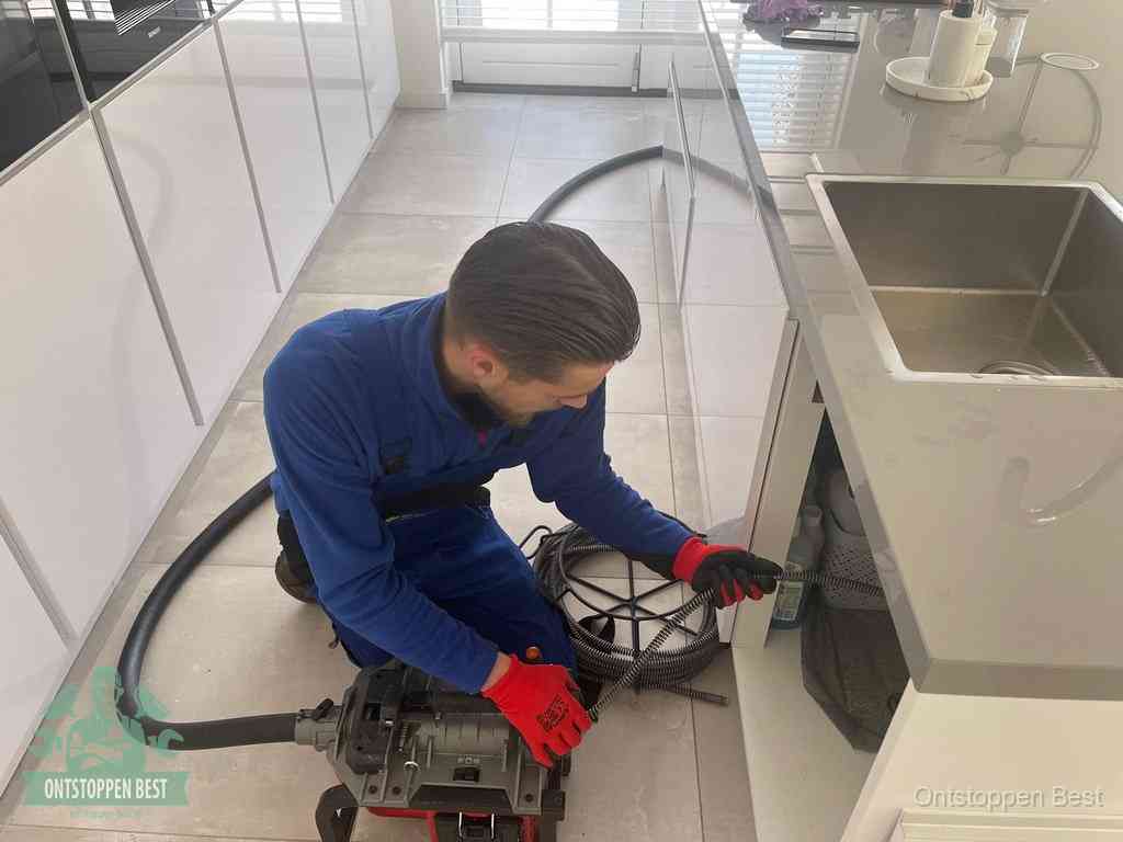 Een monteur ontstopt de keukenafvoer onder de spoelbak waarop ook de vaatwasser is aangesloten in Best